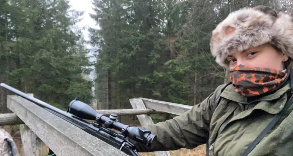 Sandra Bernal Osoba w futrzanej czapce i maskującym szaliku trzyma karabin z lunetą, stojąc na drewnianej ambonie myśliwskiej w otoczeniu gęstego lasu iglastego.