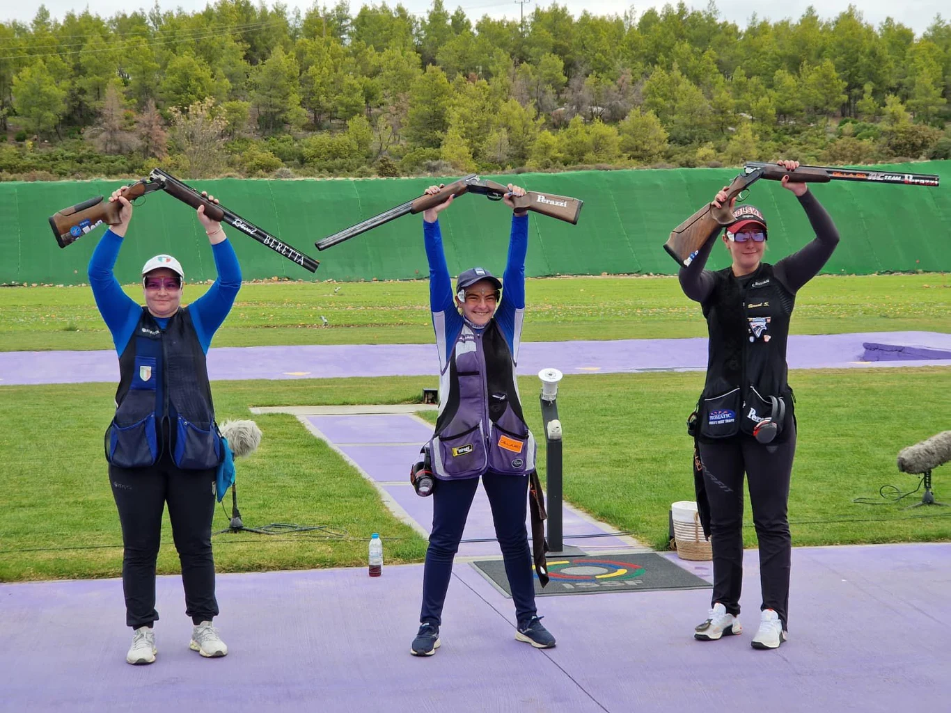 Trzy zawodniczki strzelectwa stoją na podium i unoszą nad głową swoje strzelby, każda ubrana w odzież sportową oraz kamizelkę ochronną, w tle zielony parawan i zadrzewiony teren.