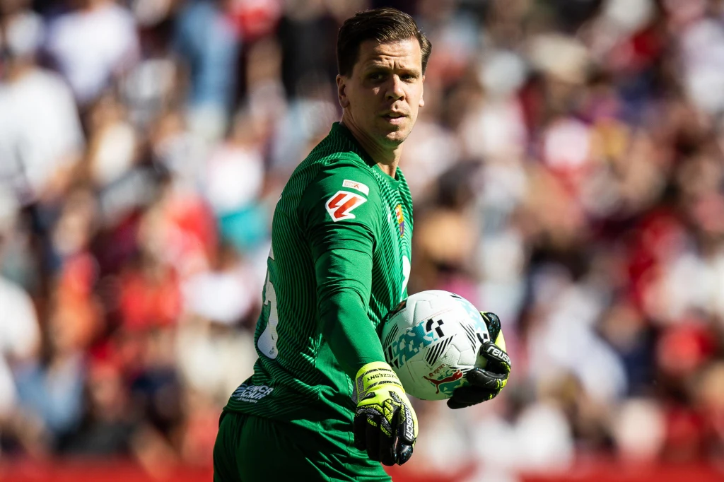 Wojciech Szczęsny Bramkarz w zielonym stroju piłkarskim trzyma piłkę w jednej ręce, skupiony na boisku, w tle rozmyty tłum kibiców na trybunach stadionu.
