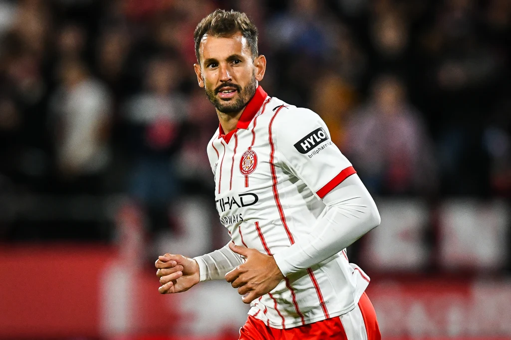 Cristhian Stuani Piłkarz w biało-czerwonym stroju sportowym z herbem klubu i logotypami sponsorów podczas meczu na stadionie, skoncentrowany na grze, w tle rozmazane sylwetki kibiców.