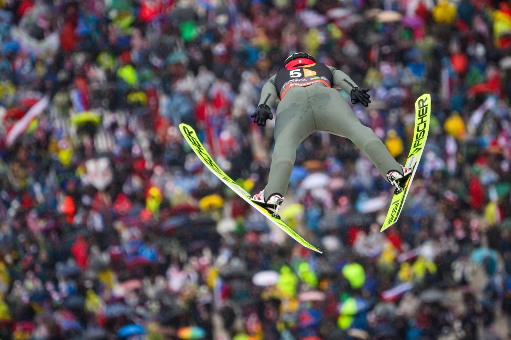 Skoczek narciarski znajdujący się w powietrzu podczas lotu na tle tłumu kolorowo ubranych kibiców.