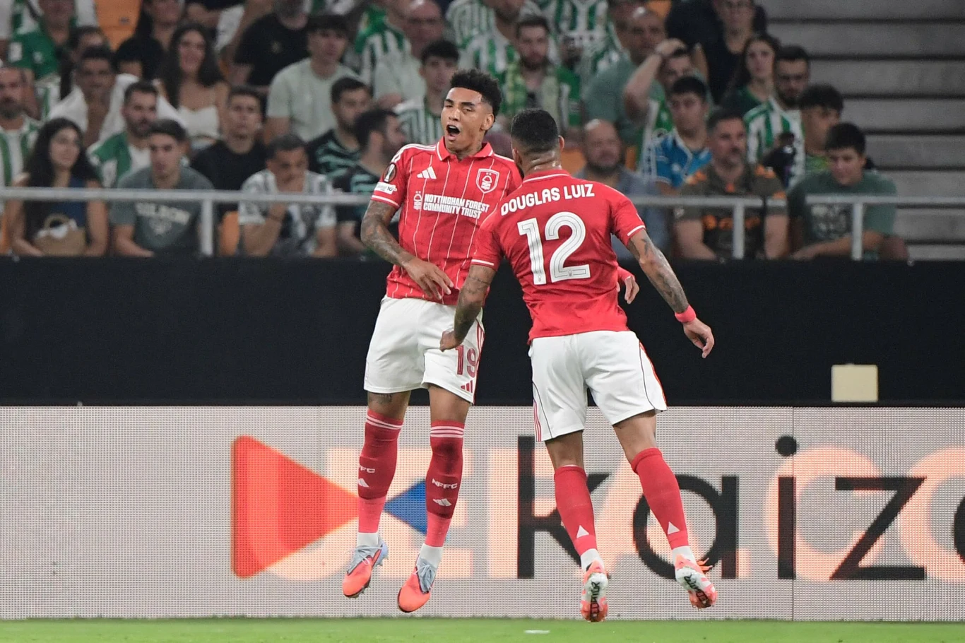 Dwóch piłkarzy drużyny w czerwonych strojach świętuje zdobycie gola na tle trybun wypełnionych kibicami, wokół widoczne są elementy stadionu i reklamy.