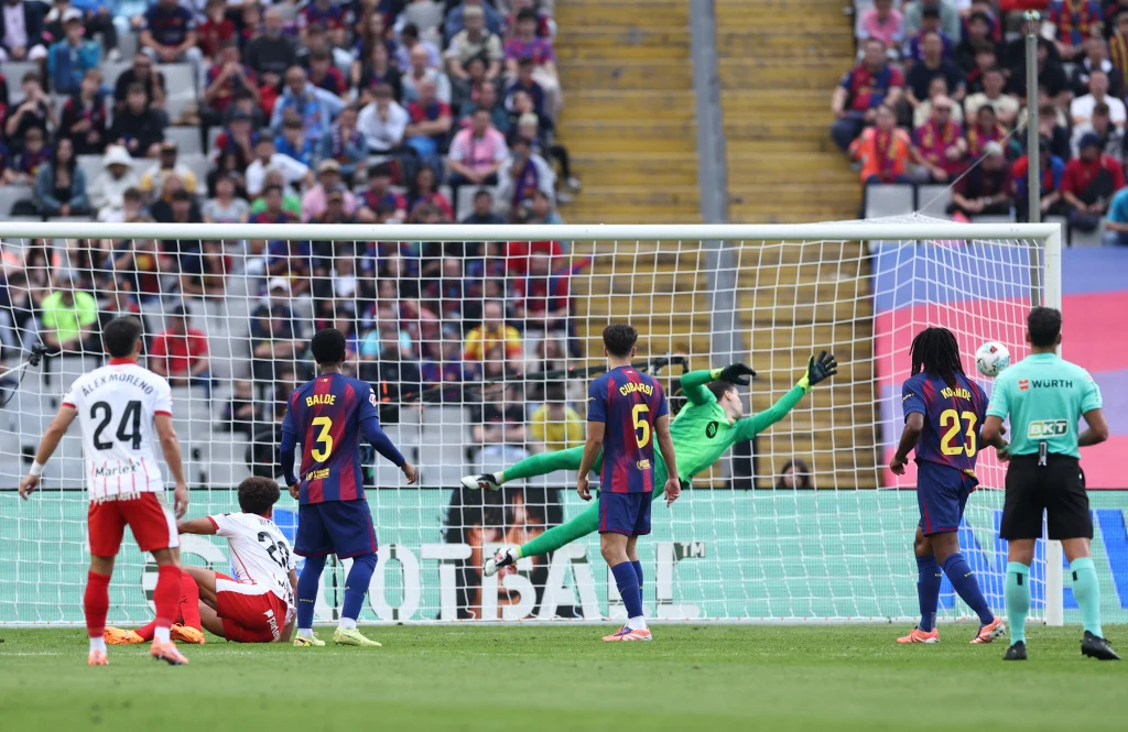 Piłkarze dwóch drużyn rywalizują podczas meczu piłki nożnej, bramkarz w zielonym stroju próbuje obronić strzał na bramkę, na stadionie widoczni kibice, sędzia obserwuje sytuację.
