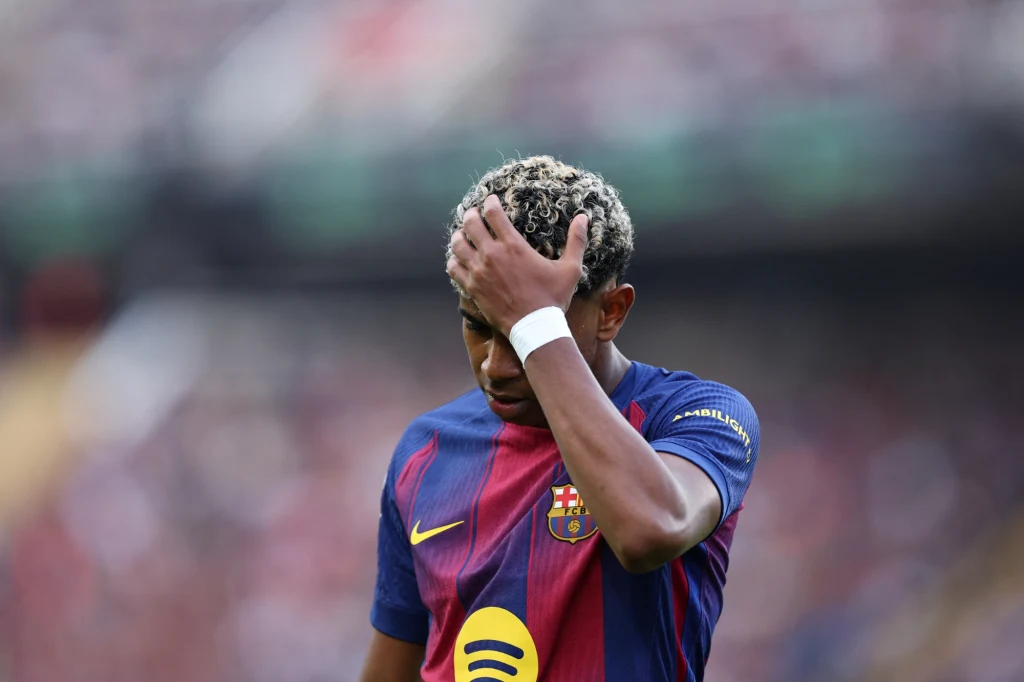 Lamine Yamal Piłkarz w koszulce FC Barcelony, z blond pasmami włosów, trzyma rękę na głowie, wyrażając zamyślenie lub zmartwienie, rozmyte tło stadionu wypełnionego publicznością.