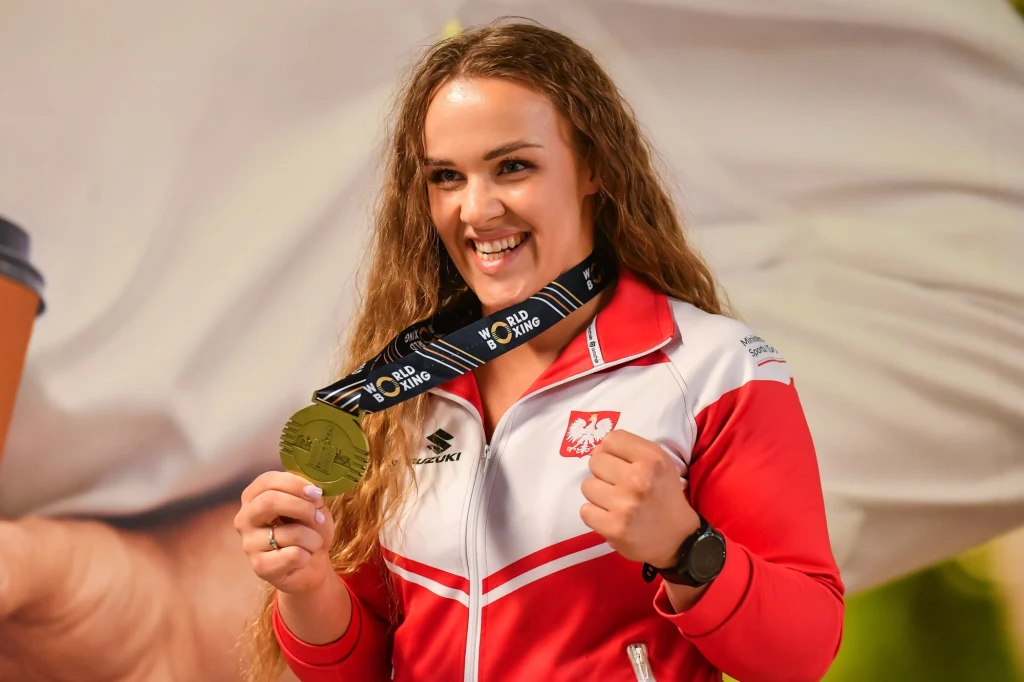Agata Kaczmarska Uśmiechnięta kobieta w stroju sportowym z orzełkiem na piersi prezentuje złoty medal zawieszony na czarnej wstążce, unosząc zaciśnięte pięści w geście radości.