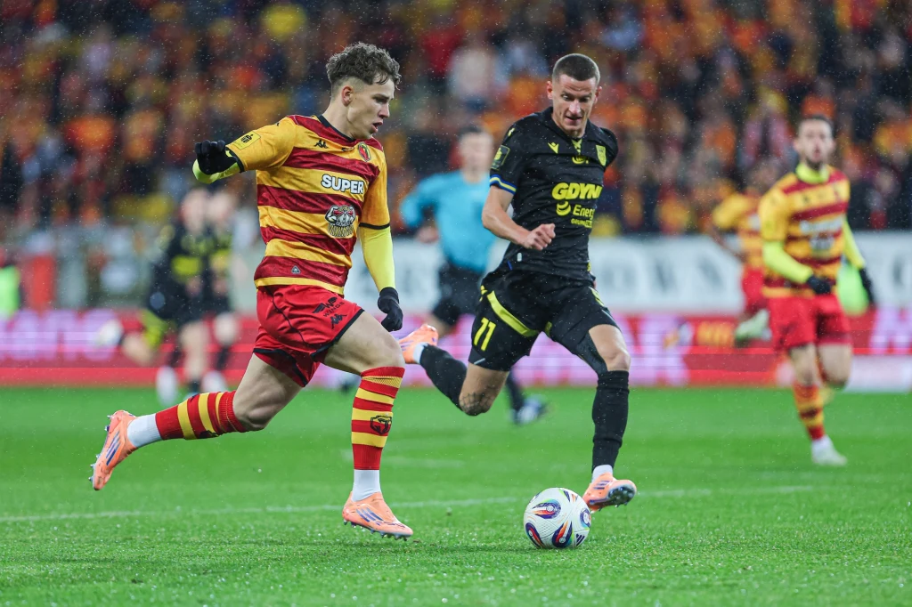 Oskar Pietuszewski Dwóch piłkarzy biegnie za piłką na boisku podczas meczu, jeden w żółto-czerwonym stroju, drugi w czarnym, wokół widownia w tle.