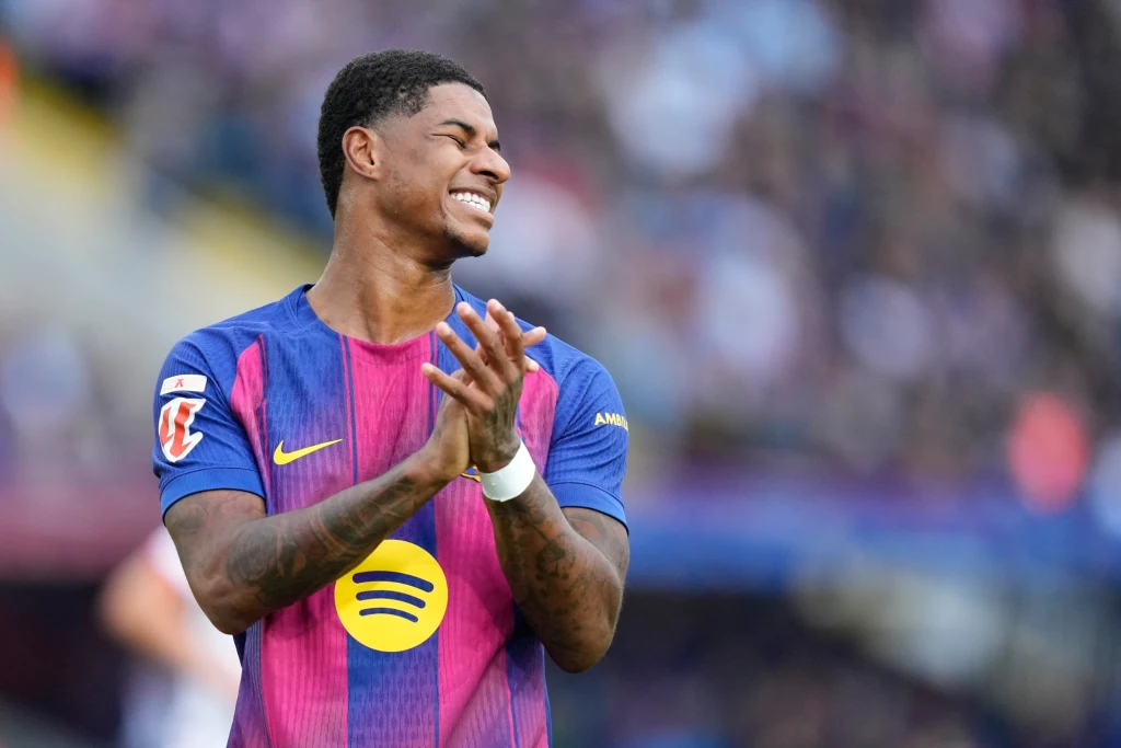 Marcus Rashford Piłkarz w fioletowo-granatowej koszulce sportowej z logo sponsora na tle stadionu, z wyrazem radości na twarzy i oklaskując rękoma