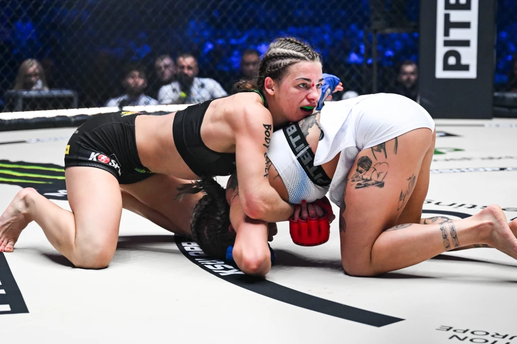 Dwie zawodniczki MMA walczą na ringu w trakcie zaciętej akcji, jedna z nich trzyma rywalkę w parterze, obie w sportowych strojach, wokół siatka oktagonu i widownia.