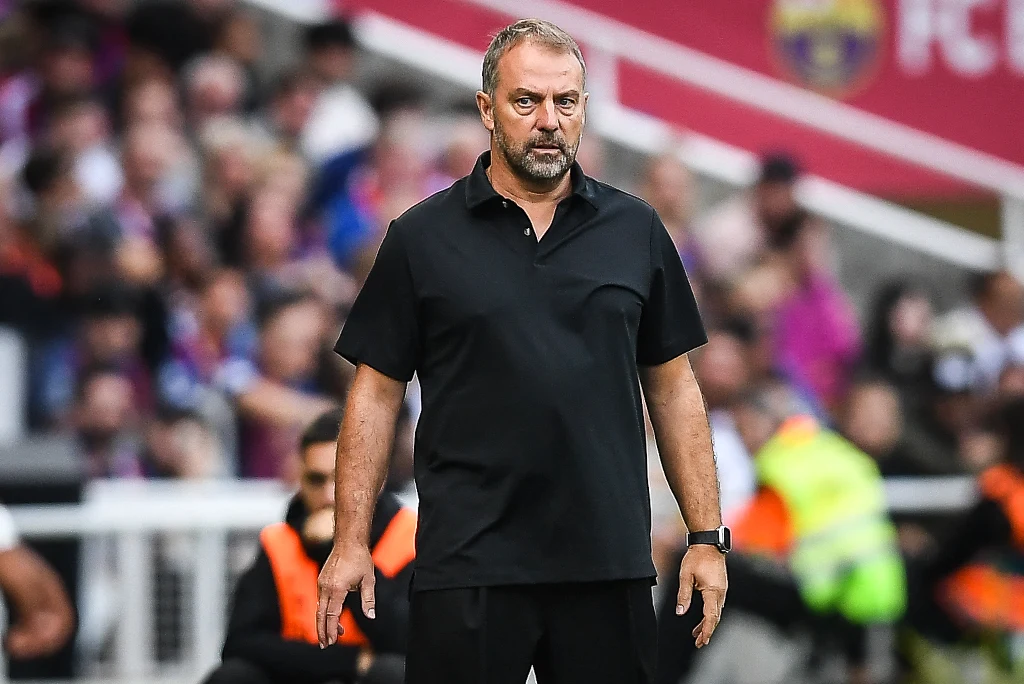 Hansi Flick Trener ubrany na czarno stoi na linii boiska podczas meczu piłkarskiego, w tle rozmyta publiczność i członkowie sztabu.