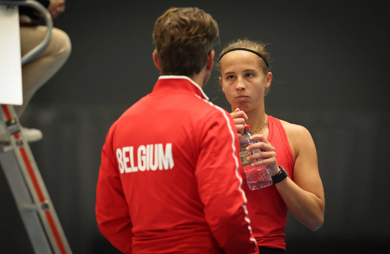 Tenisistka w czerwonym stroju pije wodę i rozmawia z trenerem w kurtce z napisem 'Belgium', w tle widoczne fragmenty sędziego siedzącego na krześle.