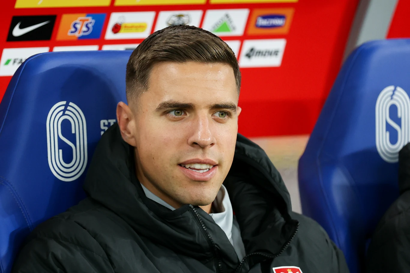 Mężczyzna w kurtce sportowej siedzący na niebieskim fotelu stadionowym, w tle widoczne tablice z logotypami sponsorów i kolorowa ścianka reklamowa.