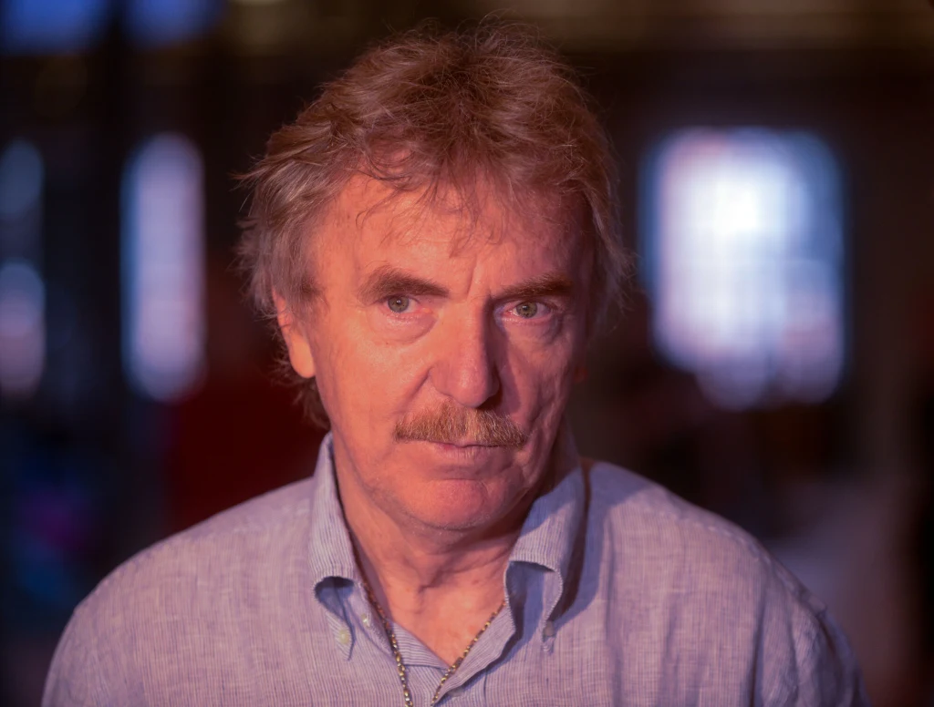 Zbigniew Boniek Mężczyzna w średnim wieku o jasnych włosach i wąsach patrzy poważnie w obiektyw, ubrany w jasnoniebieską koszulę, w tle rozmyte światła.