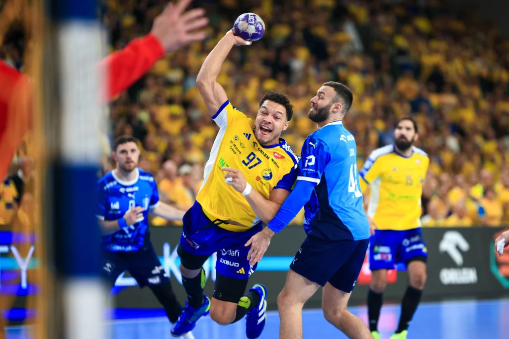 Theo Monar w akcji Intensywna akcja podczas meczu piłki ręcznej, zawodnik w żółtej koszulce, trzymając piłkę, próbuje przedrzeć się przez obronę rywala z drużyny w niebieskim stroju, w tle widoczny tłum kibiców.