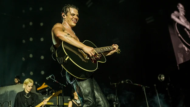 Wokalista grający na gitarze akustycznej podczas koncertu, w tle widoczny gitarzysta i scena oświetlona reflektorami, atmosfera energetycznego występu na żywo.