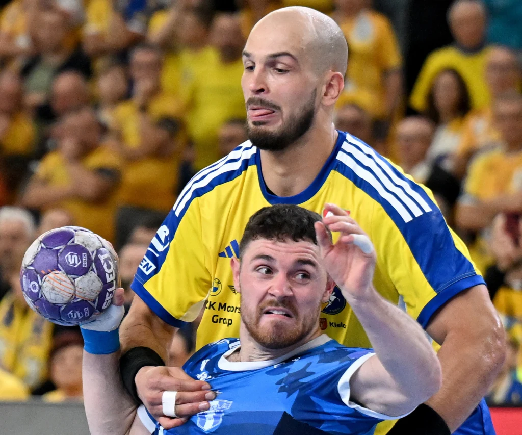 Arciom Karaliok i Mitja Janc Dwóch zawodników w trakcie intensywnej gry w piłkę ręczną, jeden ubrany w żółto-niebieską koszulkę próbuje zatrzymać przeciwnika w niebieskim stroju trzymającego piłkę, w tle widoczna publiczność.