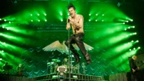 Energetyczny wokalista w skórzanych spodniach podskakuje z mikrofonem podczas występu zespołu rockowego, scena oświetlona jest intensywnie zielonymi światłami, w tle perkusja i inni muzycy, efektowne dekoracje sceniczne imitujące skrzydła dodają dramat...