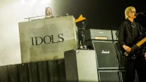 Grupa muzyków występująca na scenie, wokalista stojący przed mikrofonem oraz klawiszowiec za dużym, betonowym pulpitem z napisem IDOLS, w tle zestaw wzmacniaczy Marshall i reflektory oświetlające scenę.