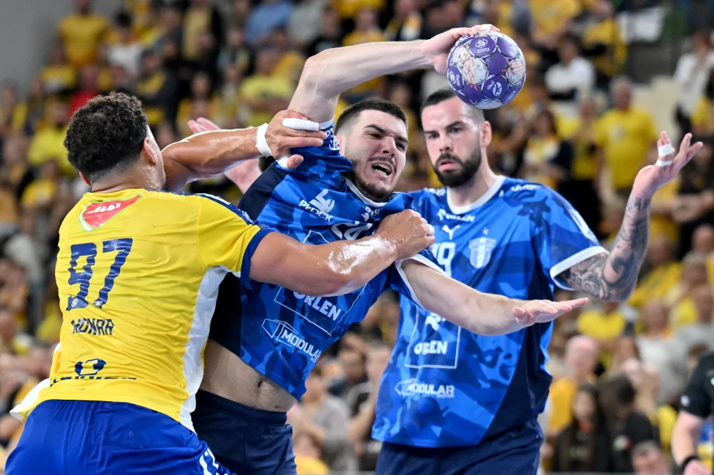 Gergo Fazekas, bohater starcia w Kielcach Intensywna akcja podczas meczu piłki ręcznej, zawodnicy drużyn w niebieskich i żółtych strojach walczą o piłkę na tle pełnych trybun kibiców.