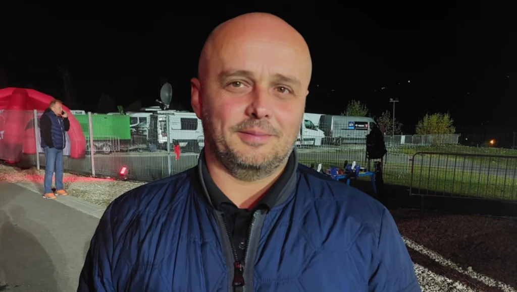 Tomasz Grzywacz - sekretarz generalny PZN łysy mężczyzna w granatowej kurtce patrzy prosto w obiektyw, z tyłu znajduje się ogrodzenie, kilka osób oraz wozy transmisyjne na tle nocnego nieba