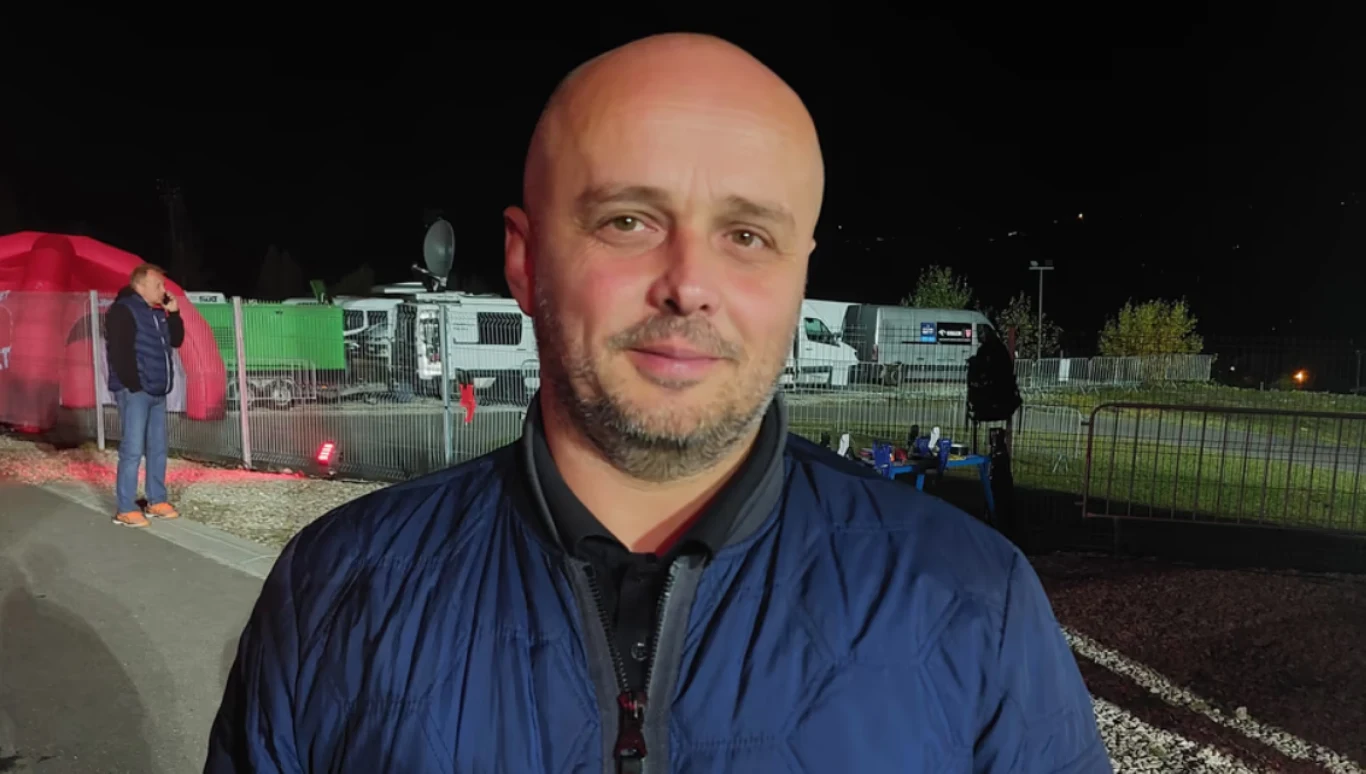 łysy mężczyzna w granatowej kurtce patrzy prosto w obiektyw, z tyłu znajduje się ogrodzenie, kilka osób oraz wozy transmisyjne na tle nocnego nieba