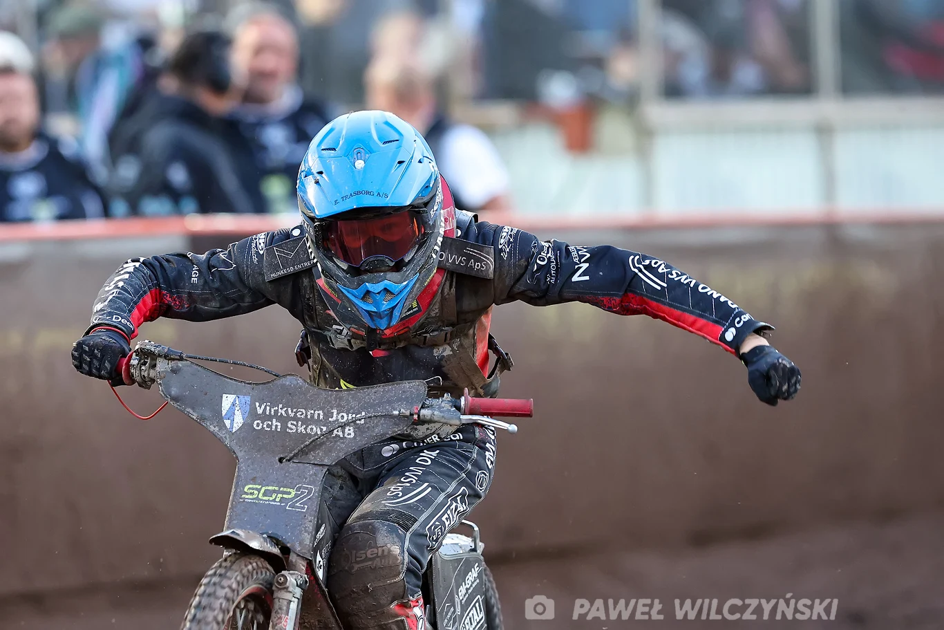 Zawodnik żużlowy w niebieskim kasku i czarnym kombinezonie w trakcie jazdy na torze, trzymający mocno kierownicę motocykla, za nim widoczne rozmyte sylwetki kibiców.