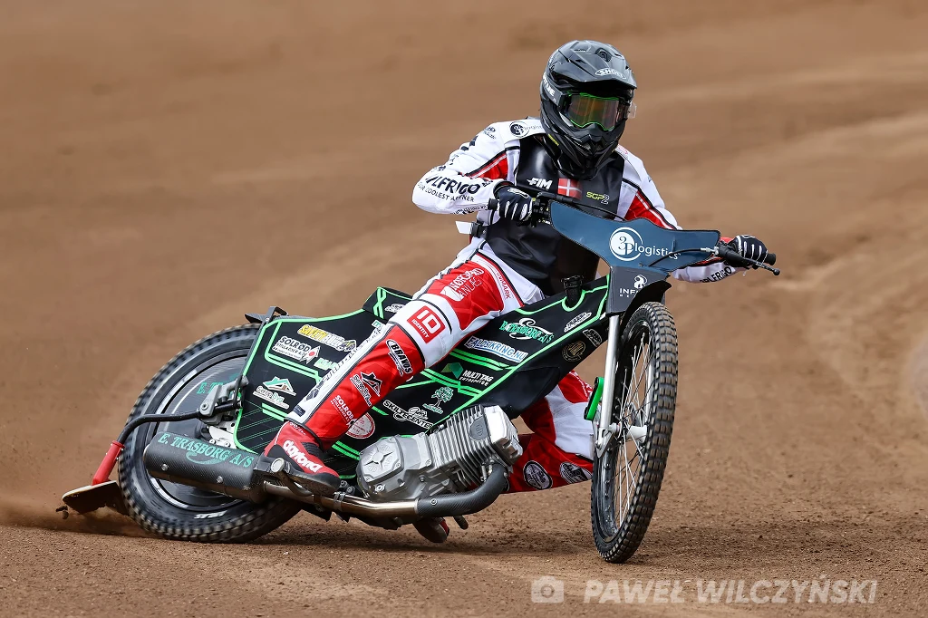 Villads Nagel w SGP2. Zawodnik żużlowy w pełnym kombinezonie i kasku pochyla się na zakręcie podczas wyścigu na torze ziemnym, prowadząc czarno-zielony motocykl żużlowy.