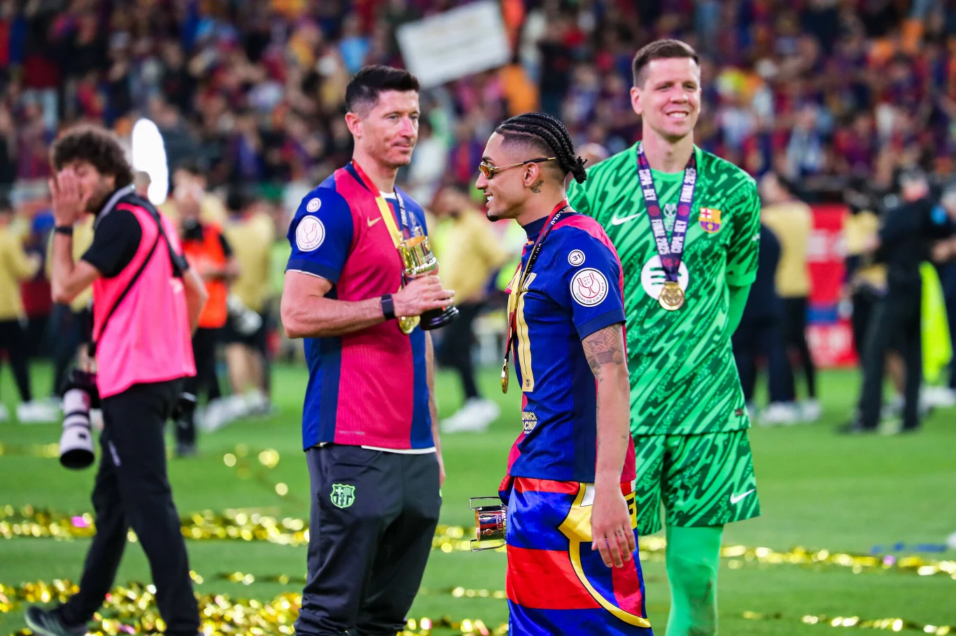 Robert Lewandowski, Raphinha i Wojciech Szczęsny Piłkarze FC Barcelony świętują zwycięstwo na boisku, otoczeni kolorowym konfetti i medalami na szyjach, w tle kibice oraz inni członkowie zespołu.