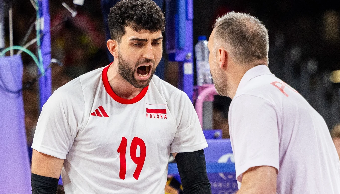 Siatkarz w białym stroju z numerem 19 wykrzykuje coś w kierunku trenera podczas meczu siatkówki, na jego koszulce widoczny napis POLSKA, w tle fragment sportowej hali.