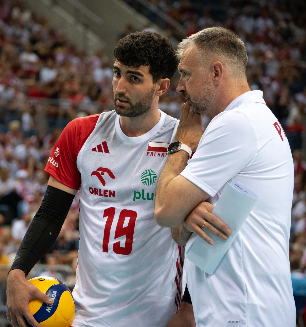 Serbski szkoleniowiec doskonale rozumiał, że Marcin Janusz potrzebuje odpoczynku Siatkarz w biało-czerwonym stroju trzymający piłkę do siatkówki rozmawia z trenerem podczas meczu, otoczeni kibicami na trybunach.