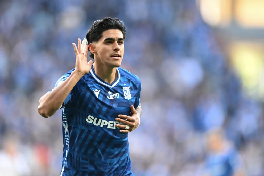 Luis Palma Piłkarz w granatowej koszulce z napisem SUPERBET świętuje zdobycie gola, przykładając dłoń do ucha i trzymając się za pierś, w tle rozmazani kibice na stadionie.