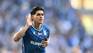 Piłkarz w granatowej koszulce z napisem SUPERBET świętuje zdobycie gola, przykładając dłoń do ucha i trzymając się za pierś, w tle rozmazani kibice na stadionie.