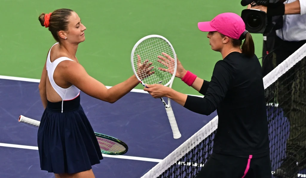 Dwie tenisistki wymieniają się uściskiem rakiet przez siatkę po zakończonym meczu na korcie, jedna z nich ubrana w czarną sukienkę, druga w ciemny strój z różowymi akcentami, w tle fragment kortu tenisowego.