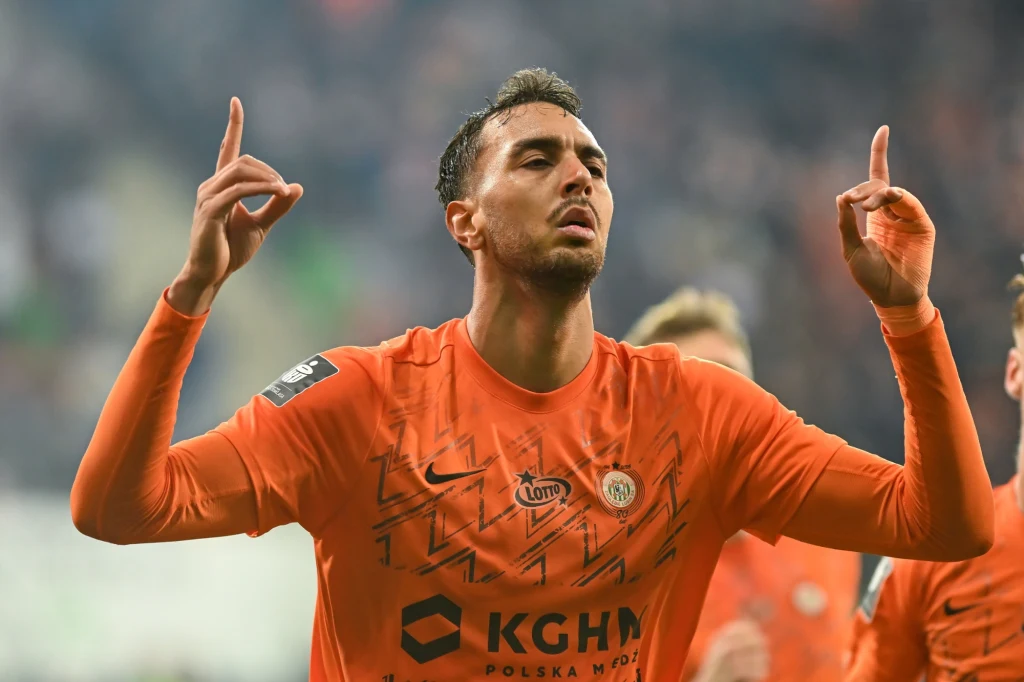 Piłkarz w pomarańczowej koszulce drużyny Zagłębie Lubin wykonuje gest z uniesionymi do góry palcami wskazującymi, w tle rozmazane sylwetki innych zawodników i kibiców na trybunach.