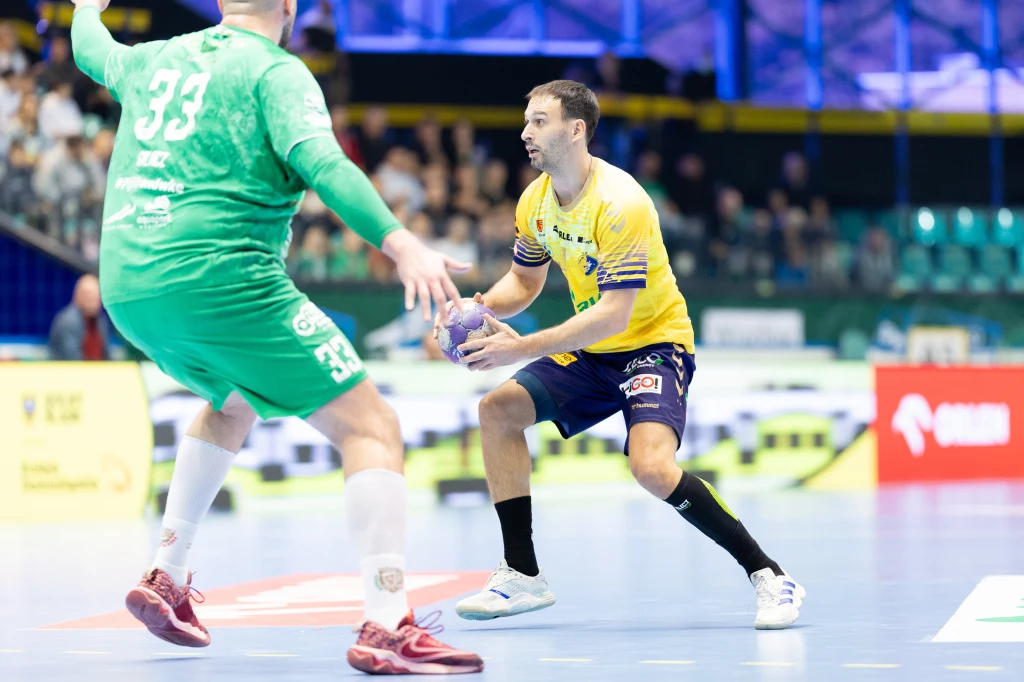 Arkadiusz Moryto Dwóch zawodników podczas meczu piłki ręcznej na hali sportowej, jeden w żółtej koszulce przygotowuje się do podania lub rzutu, drugi w zielonym stroju broni dostępu do bramki.