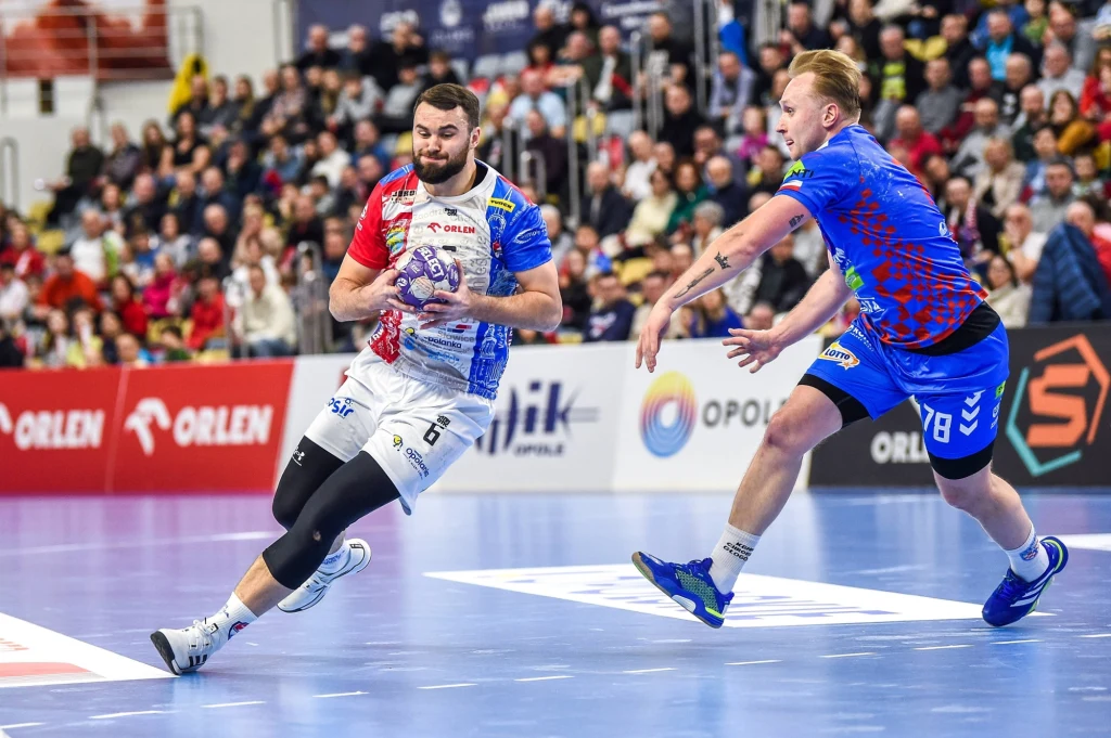 Bartosz Skiba (z prawej strony) Dwóch piłkarzy ręcznych w trakcie dynamicznej akcji na boisku sportowym, zawodnik w białym stroju prowadzi piłkę, a drugi w niebieskim stara się go powstrzymać, w tle widoczna publiczność na trybunach.