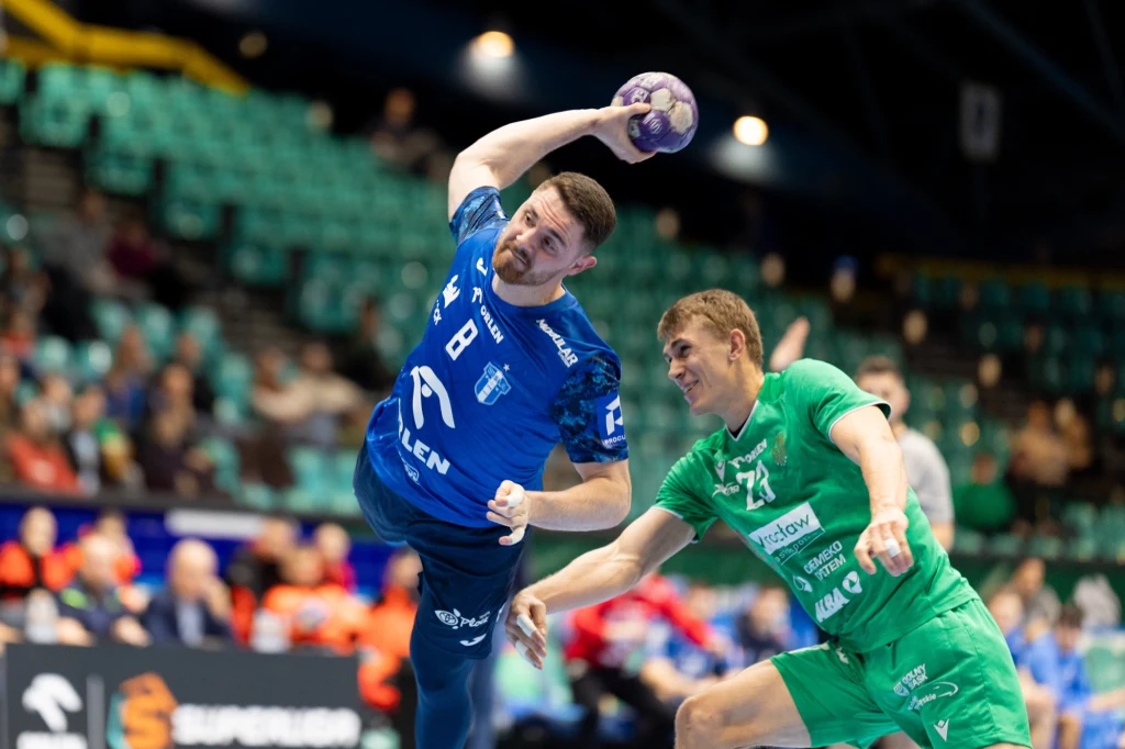 Mitja Janc (z lewej strony) Dwóch zawodników w trakcie dynamicznej akcji podczas meczu piłki ręcznej, jeden z nich ubrany w niebieski strój rzuca piłkę, drugi w zielonym stroju próbuje go zablokować, w tle widać trybuny z widzami.