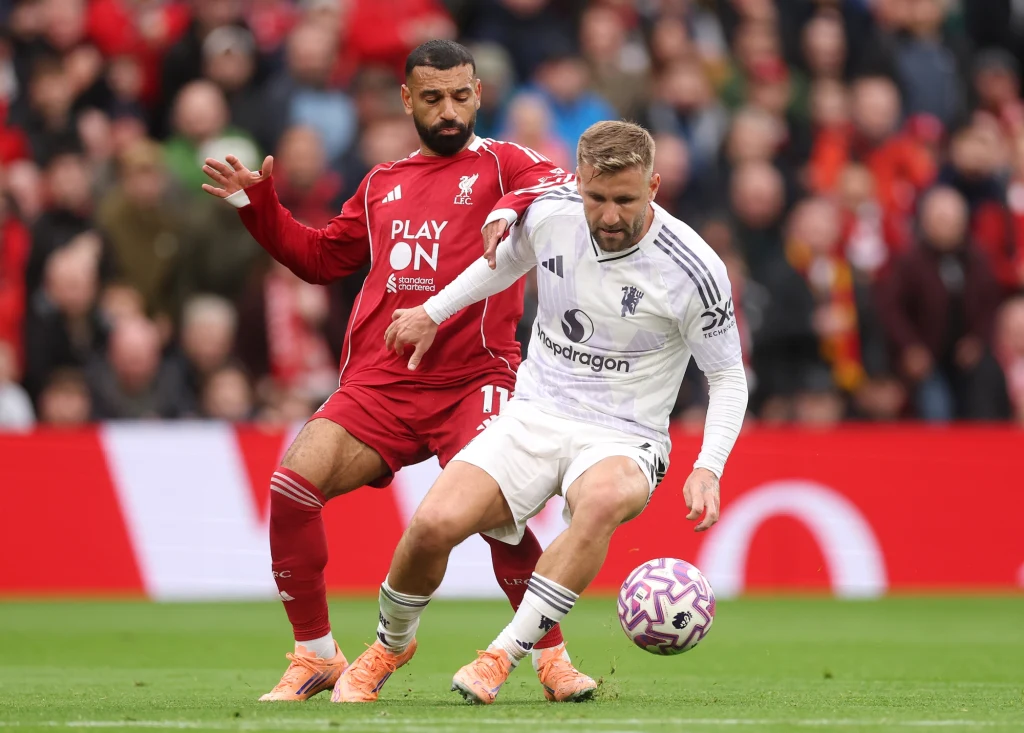 Dwóch piłkarzy walczy o piłkę na murawie podczas meczu, jeden ubrany w czerwony, drugi w biały strój, w tle widać trybuny pełne kibiców.