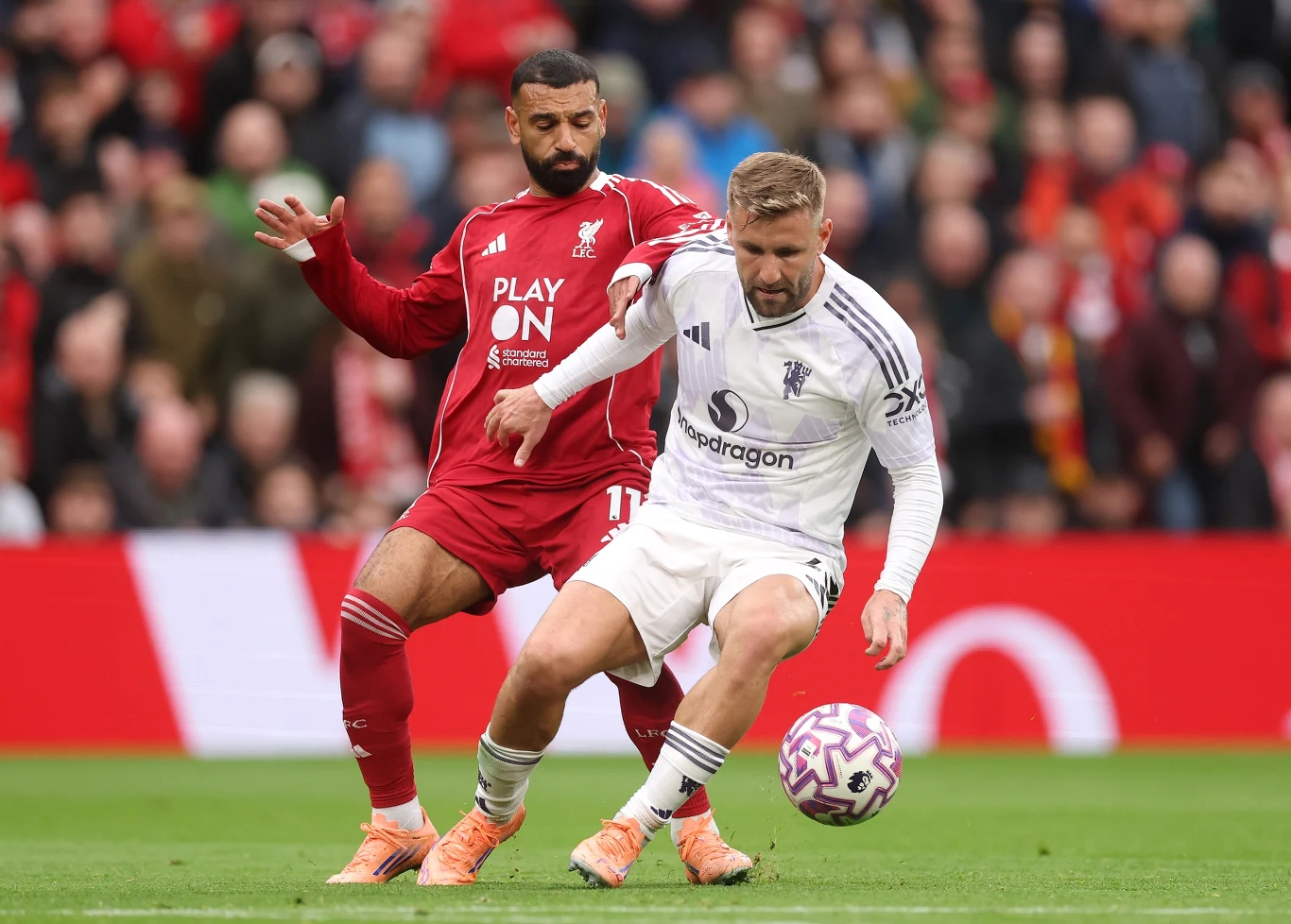 Dwóch piłkarzy walczy o piłkę na murawie podczas meczu, jeden ubrany w czerwony, drugi w biały strój, w tle widać trybuny pełne kibiców.