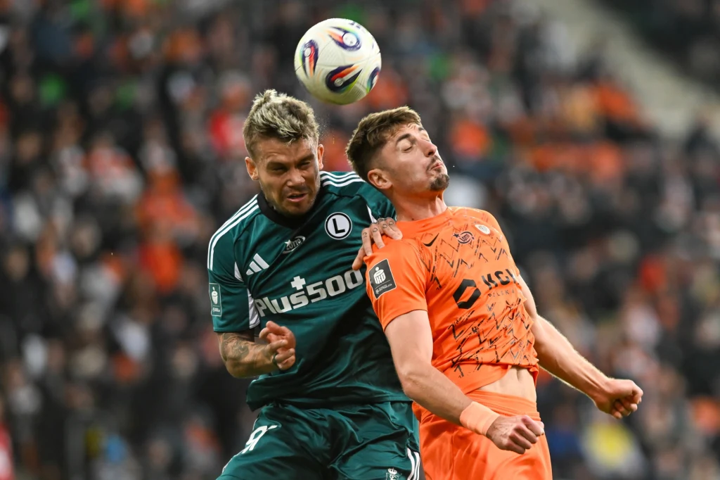 Dwóch piłkarzy rywalizuje o piłkę w powietrzu, jeden ubrany w zielony, drugi w pomarańczowy strój, wykonując główkę w trakcie meczu na stadionie z zamazaną publicznością w tle.