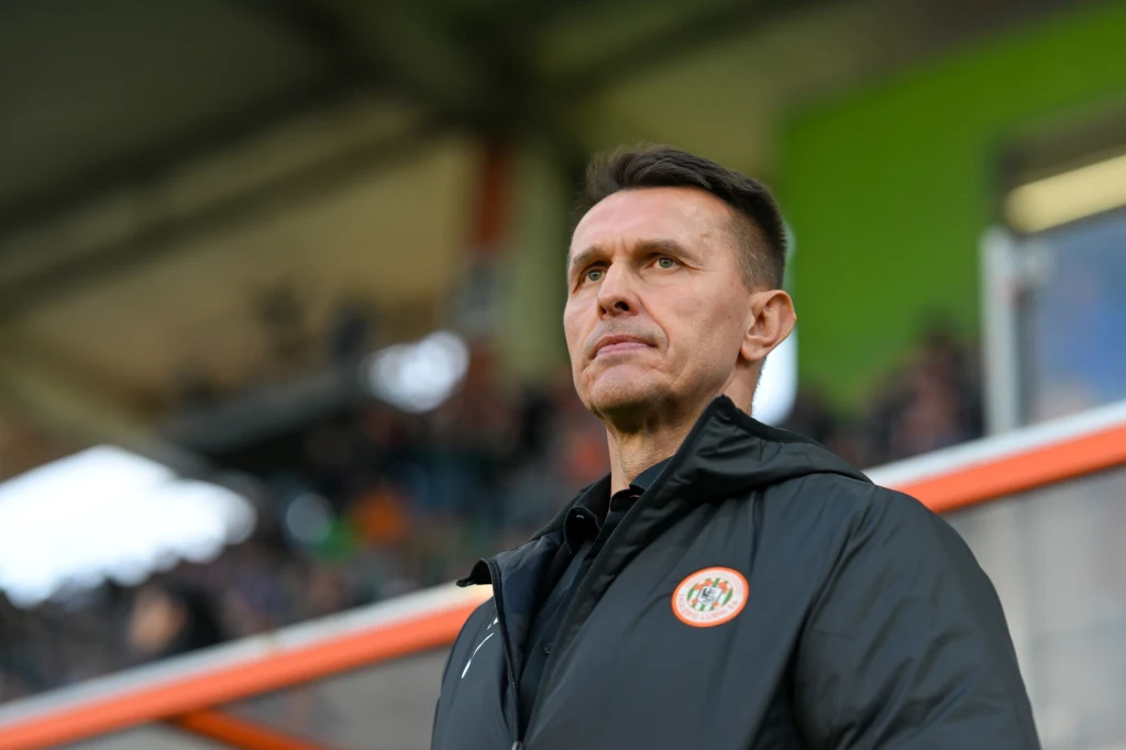 Mężczyzna w czarnej kurtce z herbem piłkarskiego klubu stoi na tle trybun stadionu, patrząc poważnie przed siebie