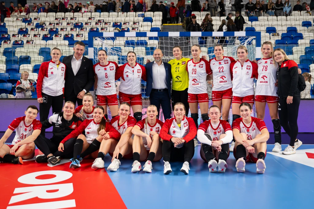 Grupa kobiet w strojach sportowych do piłki ręcznej wraz ze sztabem trenerskim pozuje na hali sportowej, większość drużyny klęczy lub siedzi na parkiecie przy linii bocznej, w tle widać trybuny z widownią.