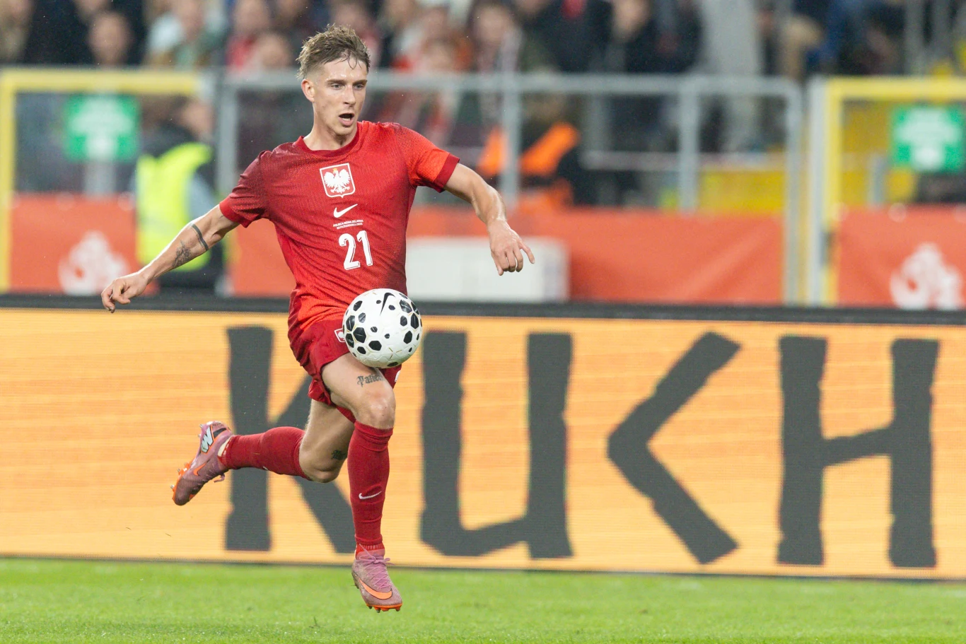 Michał Skóraś Polski piłkarz w czerwonym stroju z numerem 21 kontroluje piłkę na boisku podczas meczu, w tle widoczna jest rozmyta publiczność oraz reklama na stadionie.