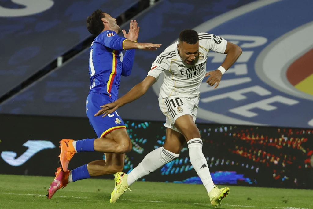 Dwóch piłkarzy w dynamicznej walce o piłkę na murawie, jeden z nich w stroju białym wykonuje gwałtowny ruch ręką, drugi w niebieskim stroju chwieje się do tyłu po kontakcie fizycznym podczas meczu.