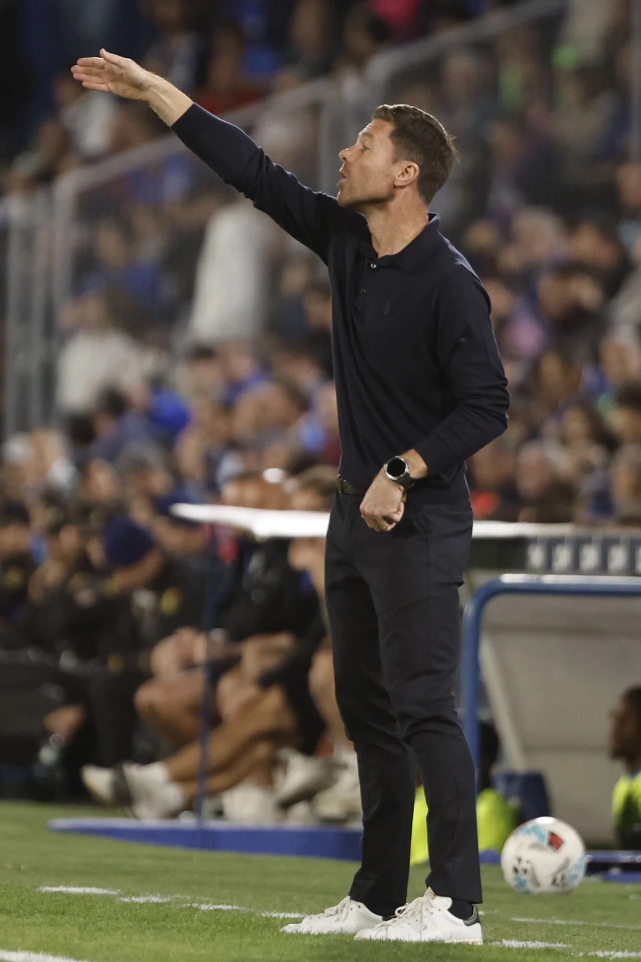 Trener ubrany w ciemny strój sportowy gestykuluje przy linii bocznej boiska podczas meczu piłkarskiego, w tle rozmyta publiczność oraz piłkarze siedzący na ławce rezerwowych.