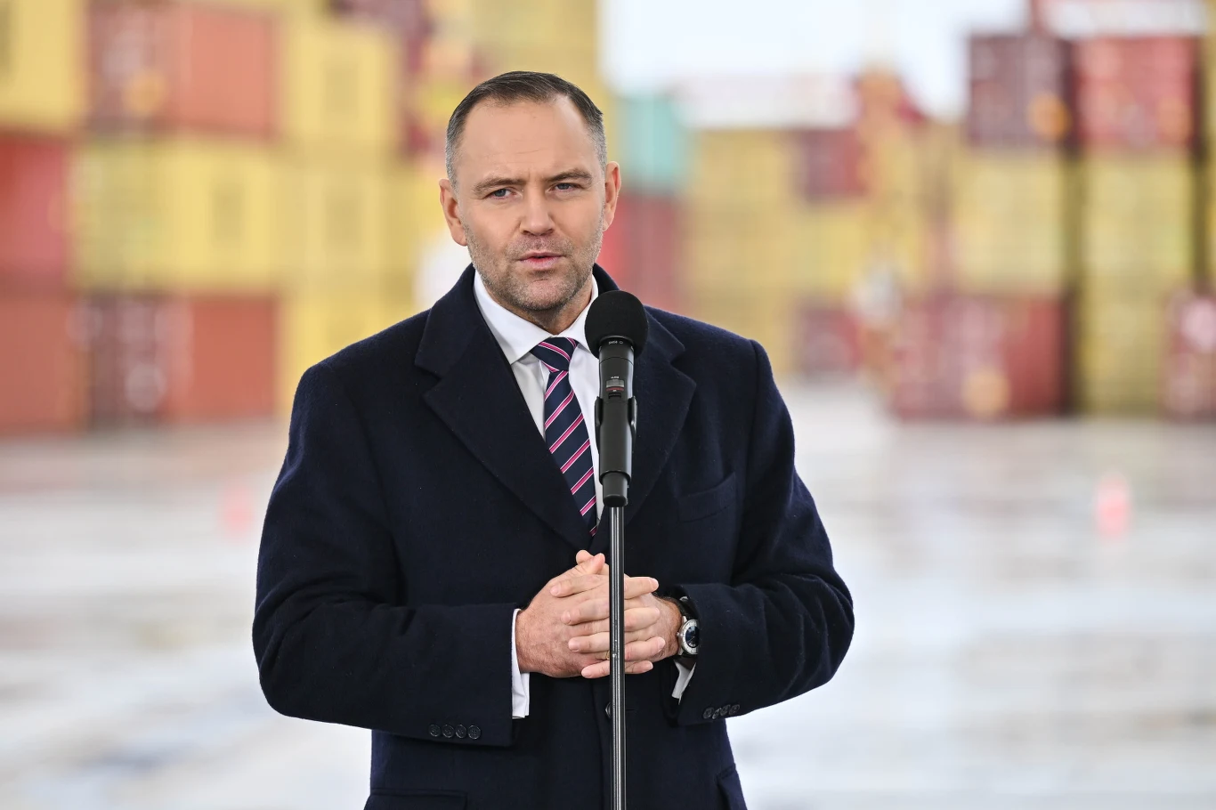 Karol Nawrocki powinien powołać własną partię? Nowy sondaż Mężczyzna w garniturze i płaszczu przemawia do mikrofonu na tle ustawionych w stosy kontenerów transportowych.