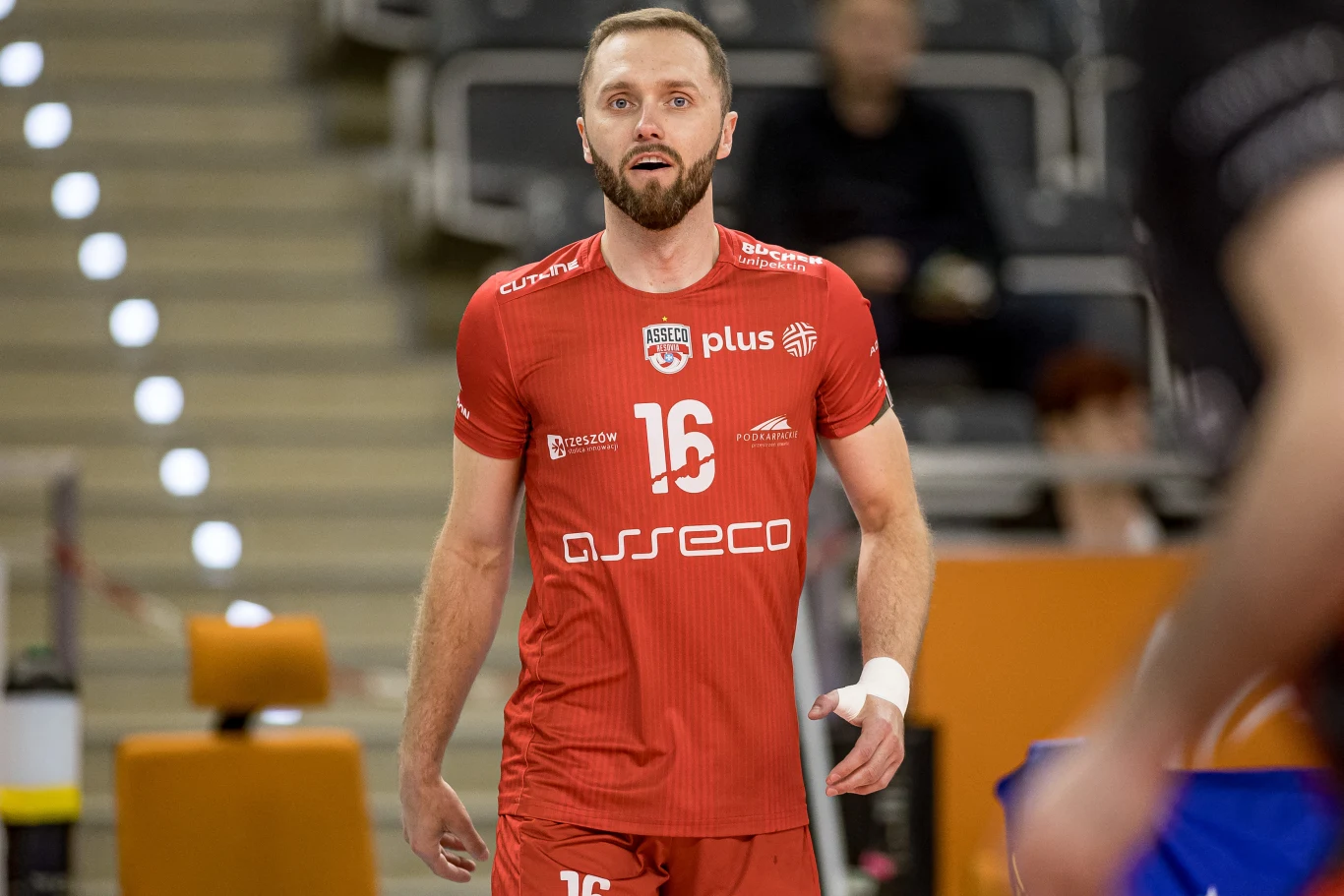 Siatkarz w czerwonym stroju drużyny Asseco Resovia z numerem 16 na koszulce znajduje się na hali sportowej, za nim widoczne są trybuny i rozmyci kibice.