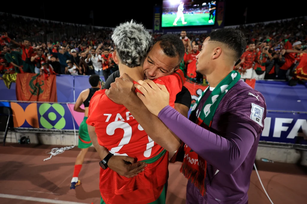 Trzech piłkarzy w strojach reprezentacyjnych obejmuje się na płycie stadionu, radośnie świętując zwycięstwo, w tle tłum kibiców z flagami.
