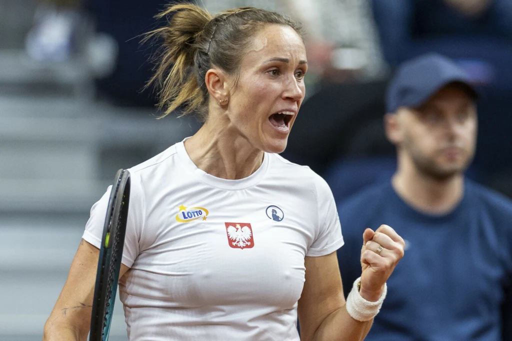 Katarzyna Kawa Tenisistka w białej koszulce i z widocznym logotypem Lotto energicznie okazuje emocje, zaciśnięta pięść sugeruje świętowanie zdobytego punktu podczas meczu, w tle widoczna rozmyta postać mężczyzny.