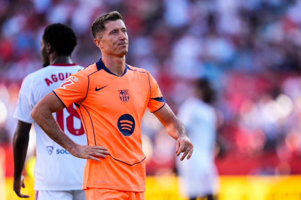 Robert Lewandowski Piłkarz w pomarańczowym stroju FC Barcelony stoi na murawie z rękami na biodrach, w tle rozmyci zawodnicy przeciwnej drużyny w białych strojach oraz trybuny stadionu.