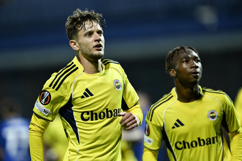 Sebastian Szymański Dwóch piłkarzy w żółtych strojach sportowych z logo klubu i napisem sponsorującym, jeden z nich ma jasne włosy i zarost, drugi ciemniejszą karnację i krótsze włosy, znajdują się na boisku i są skupieni na grze.